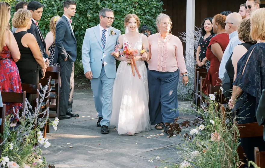A bride in a long veil walks down a flagstone aisle between both of her parents, guests standing on either side