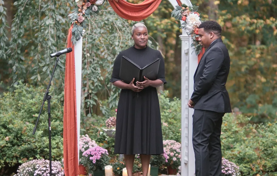 An officiant holds an open binder beneath a flower-draped garden arch while the groom stands beside her waiting