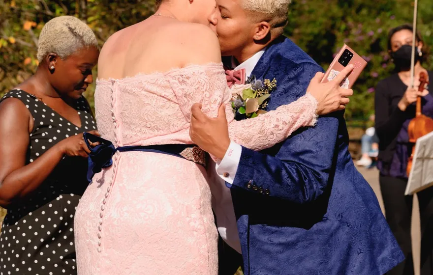 Two spouses embrace and kiss on the cheek at a vow renewal by the water, the officiant beside them and a violinist playing in the background