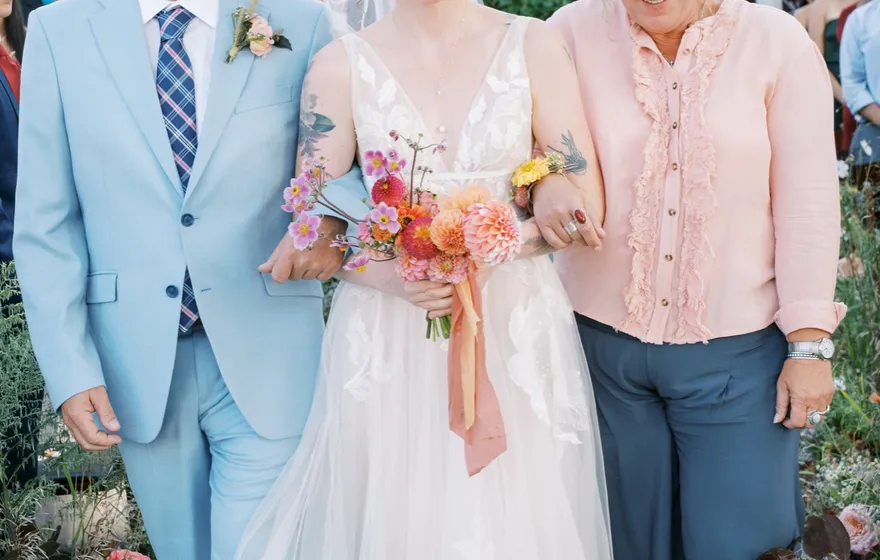 A bride walks down a garden aisle between a man in a light blue suit and a woman in a pink cardigan, guests standing on both sides