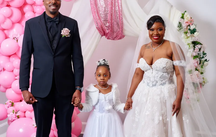 A groom in a black suit and a bride in an off-the-shoulder gown stand on either side of a young girl in a white dress, pink balloons behind them