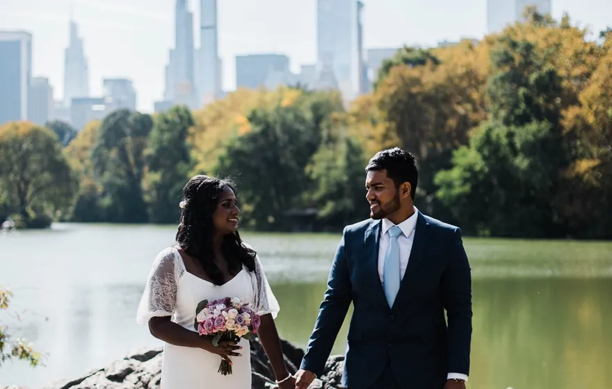 A bride and groom hold hands on the rocks at the edge of the Lake in Central Park, autumn trees and Midtown towers behind them