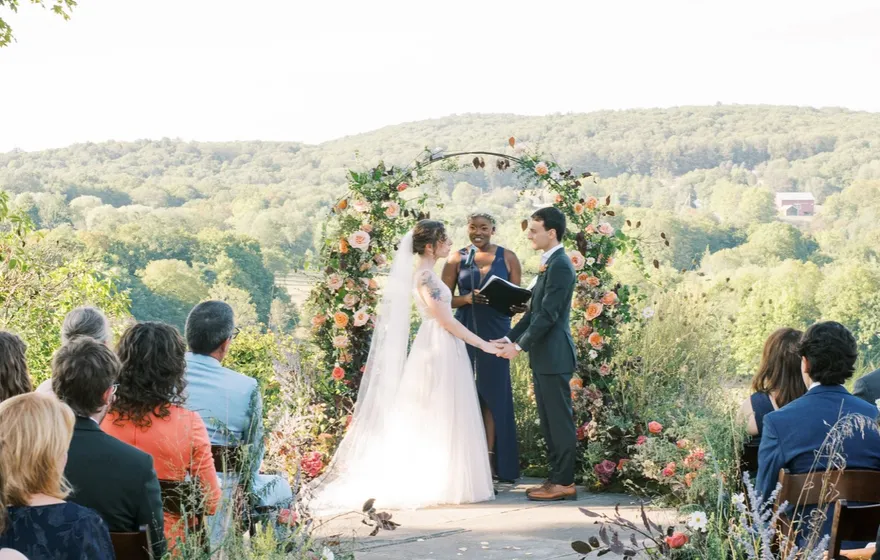 Looking down a stone aisle lined with flowers toward a couple and their officiant under a floral arch, a few rows of guests seated on either side and hills beyond