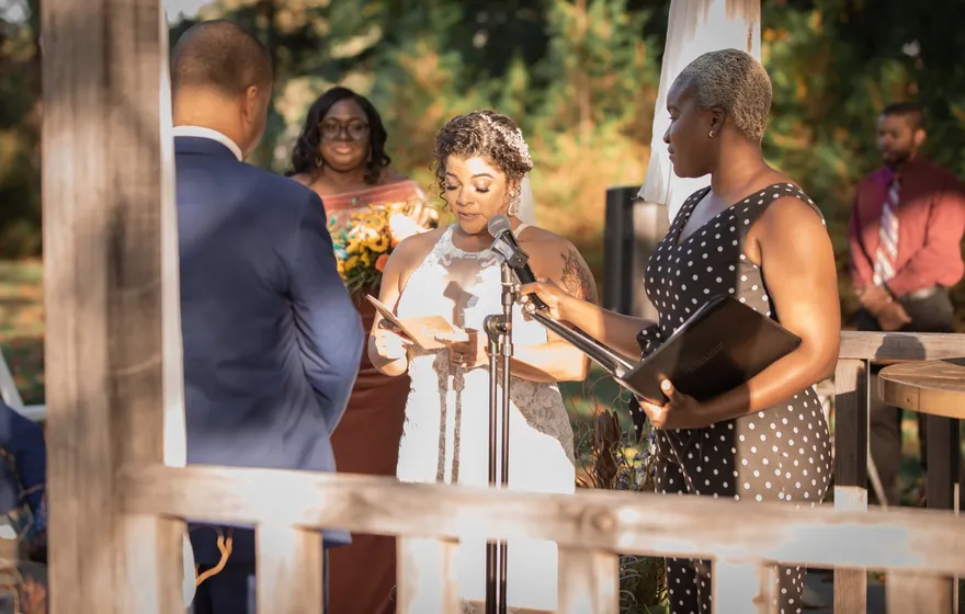 A bride reads from a card at a microphone stand while the officiant holds her binder beside her and guests look on