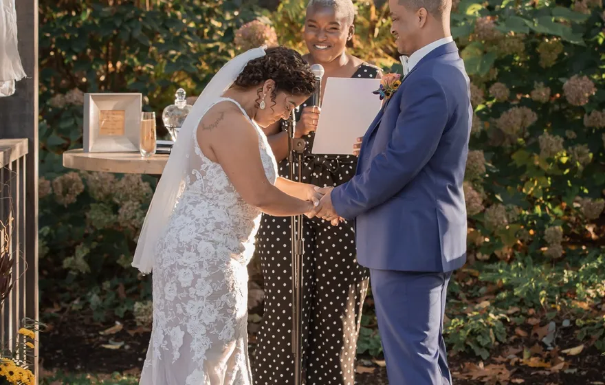 A bride and groom bow their heads together under a draped wooden arbor while the officiant behind them holds her printed script