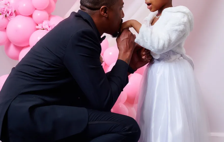 A groom in a dark suit crouches to kiss the hand of a little girl in a white dress and tiara in front of a pink balloon arch