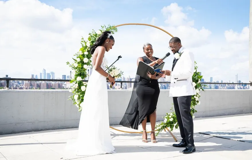 On a rooftop above the river, the officiant speaks into a microphone from her binder while a bride and groom in white stand under a round floral arch