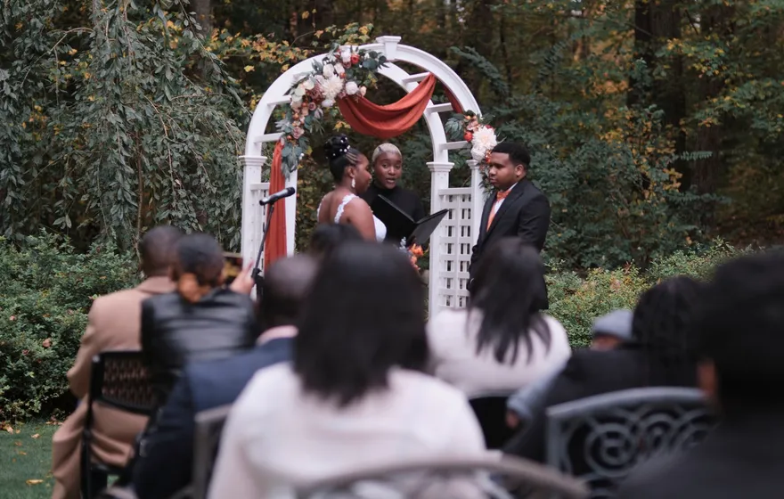 Guests seated in the foreground watch a couple and their officiant at a white arch draped in rust fabric in a wooded garden