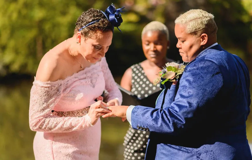 A woman in a pink lace dress places a ring on her spouse's finger while the officiant watches from behind