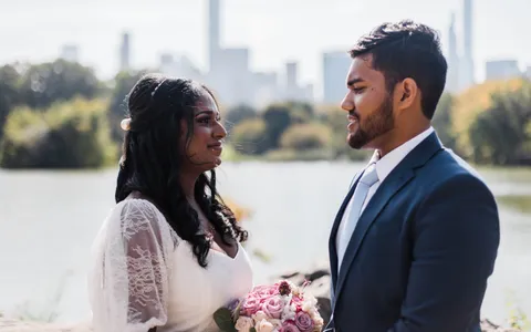 A bride holding a pink bouquet and a groom in a navy suit smile at each other beside the lake in Central Park, the Midtown skyline behind them