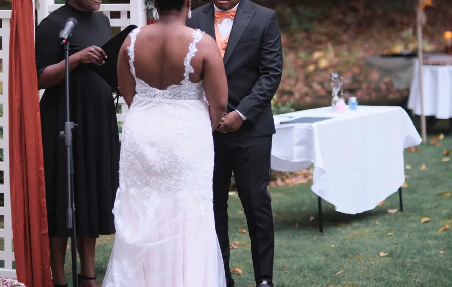 The officiant reads into a microphone at a garden arch while the bride, seen from behind, and the groom listen