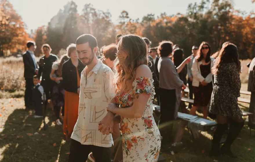 A newly married couple holds hands and smiles as guests gather around them in a sunlit field