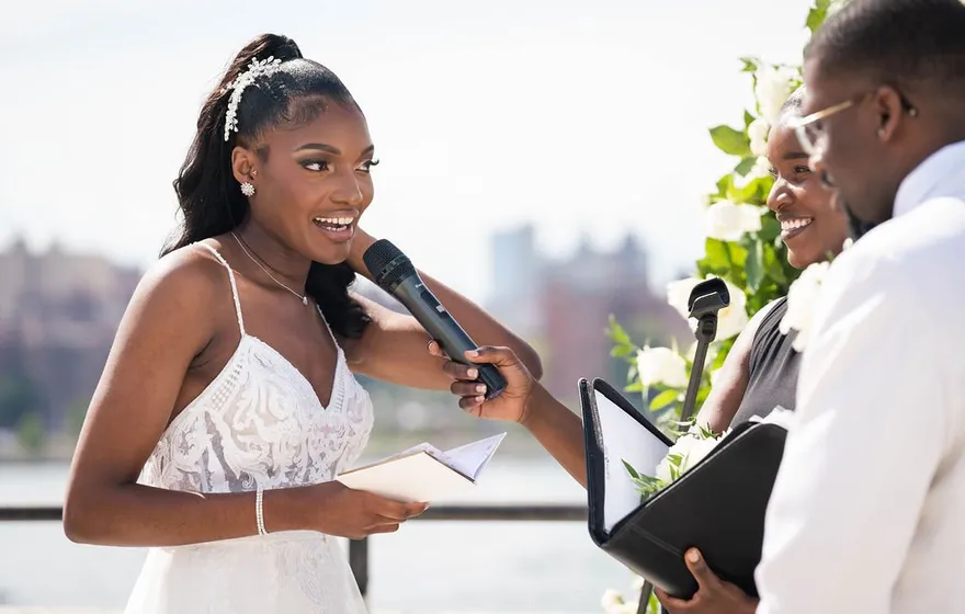 A bride holds a microphone and reads from a sheet of paper on a rooftop while the officiant beside her holds an open folder