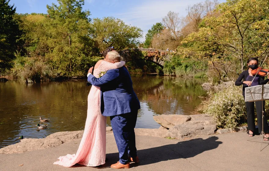 A couple embraces on the rocks beside the pond near Gapstow Bridge while a violinist plays from a music stand a few steps away