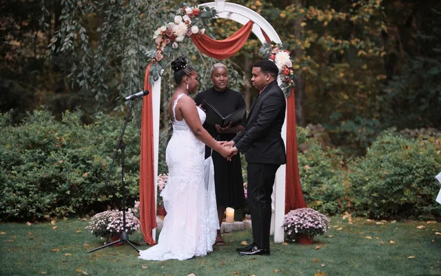A couple holds hands beneath a white garden arch draped in rust fabric while the officiant reads from an open binder