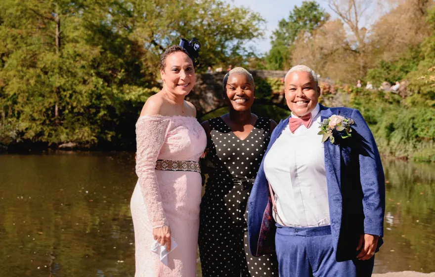 A couple renewing their vows stand with their officiant on the rocks by the Pond in Central Park, one in a long pink lace gown and one in a navy suit