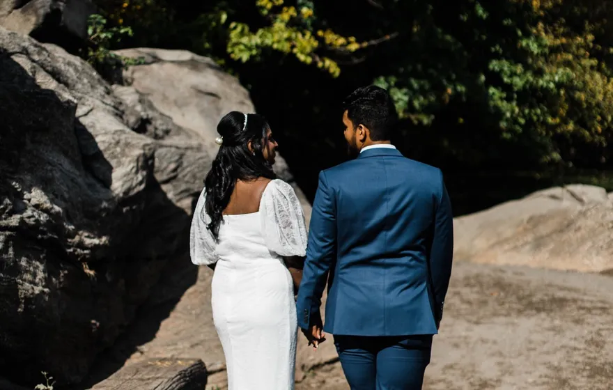 A bride and groom walk away hand in hand along a rocky path by the Lake in Central Park