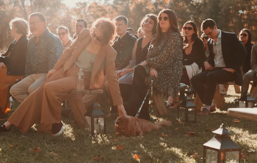 Guests seated on wooden benches at an outdoor fall ceremony, low sun lighting their faces, a woman shading her eyes as a small dog sits at her feet