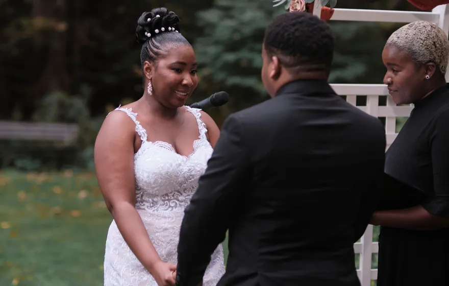 A bride speaks into a microphone while holding her groom's hands, the officiant standing close beside them in a garden