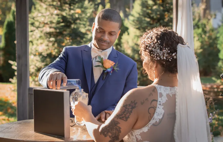 A groom pours into a glass carafe at a small round table beside a wooden box while his bride holds the carafe steady