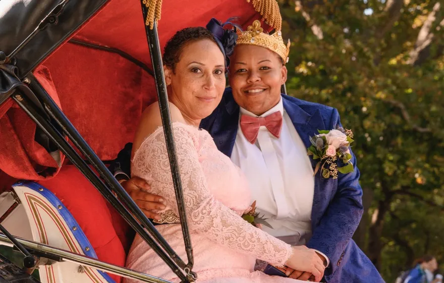 A renewing couple, one in a pink lace gown and one in a paper crown and bow tie, sit together in a red-cushioned horse carriage in Central Park