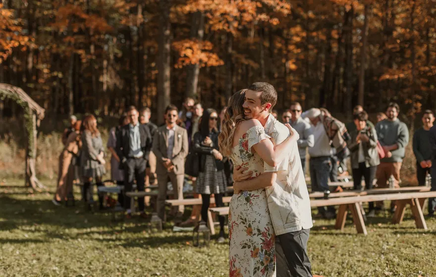 A newly married couple hug in front of the wooden benches as their guests stand and applaud in an autumn field