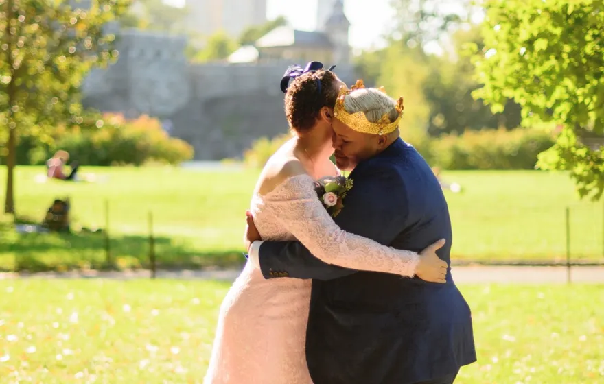 A couple in a pink lace gown and a blue suit hug on a sunny lawn, trees and stone towers behind them