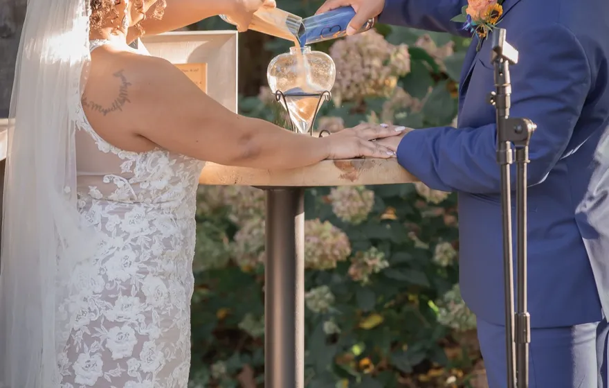 A bride and groom pouring together into a glass vessel on a small ceremony table in a garden