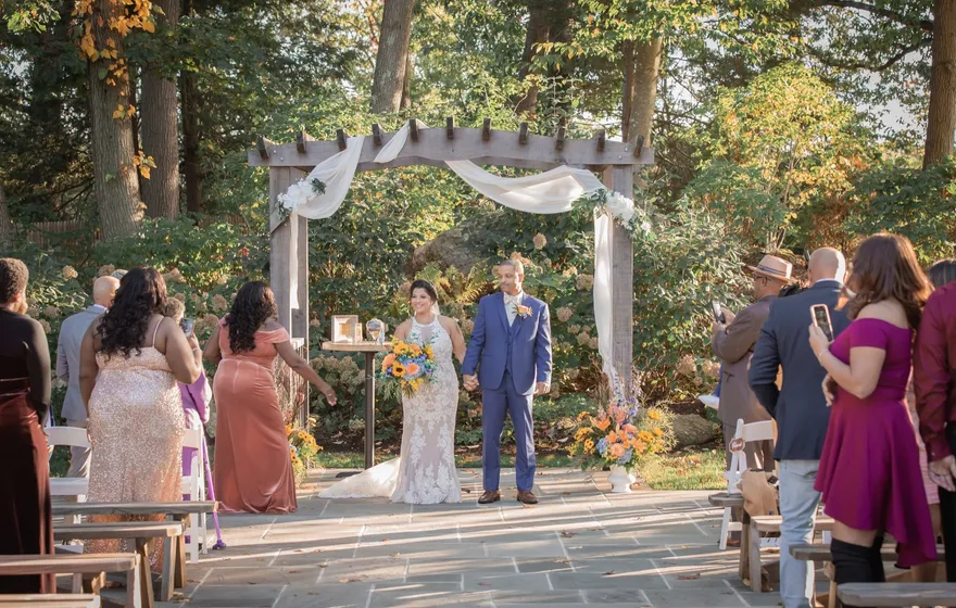 A couple holding hands under a wooden arch draped in white fabric in a leafy garden, guests standing along the aisle