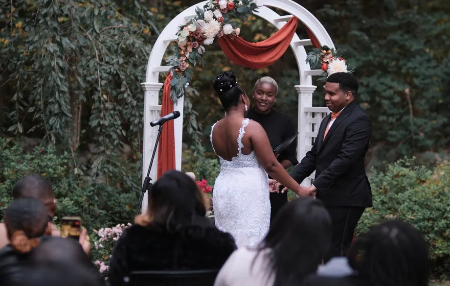 A wedding ceremony seen from behind the seated guests: the couple holds hands under a white arch draped in rust fabric while the officiant faces them