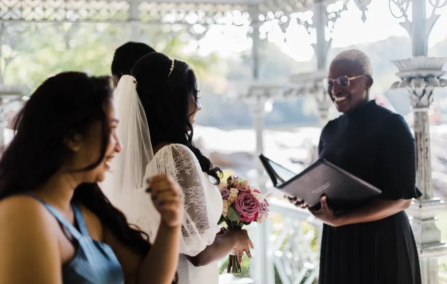 An officiant in black reads from an open folder to a bride in a lace veil under the white iron canopy of the Ladies' Pavilion in Central Park