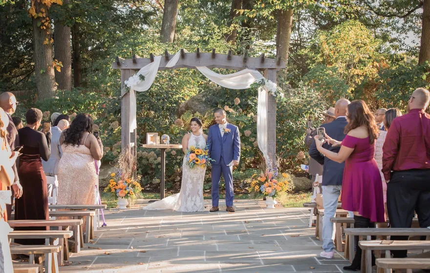 Guests standing on both sides of a stone aisle, some holding up phones, as a newly married couple stands together under a draped garden arbor