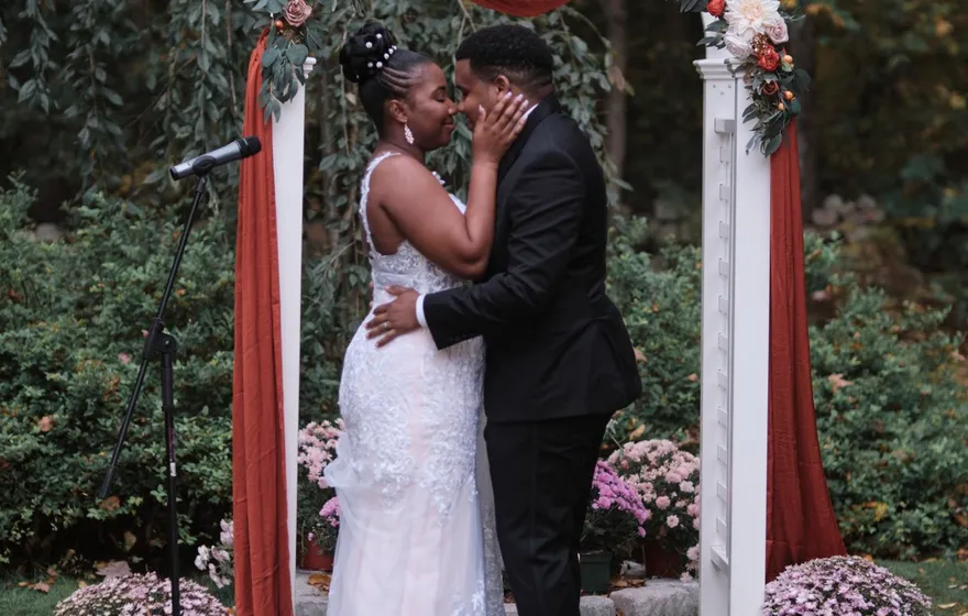 A bride and groom kiss under a white garden arch draped in rust fabric, a microphone stand beside them