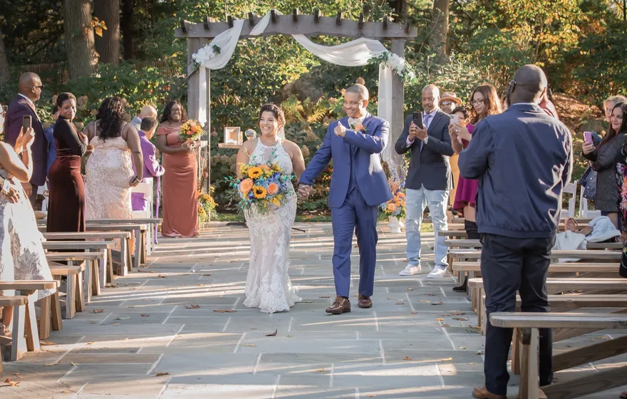 A couple walks back up a stone aisle holding hands while guests stand along wooden benches, several taking photos on their phones