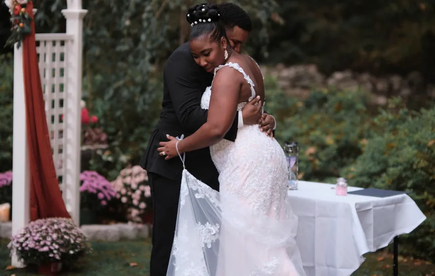 A bride and groom hold each other in a long embrace beside a white garden arch