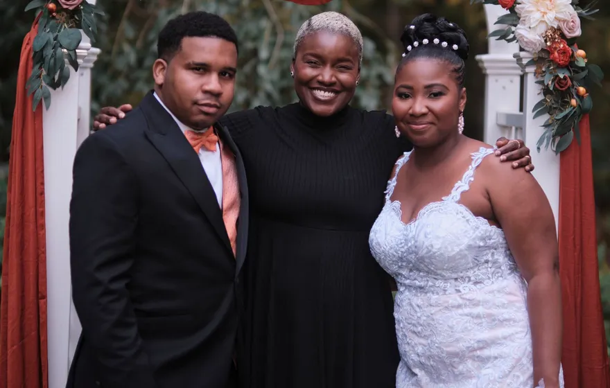 The officiant stands smiling between a groom in a black tuxedo and a bride in a lace gown in front of a floral arch after the ceremony