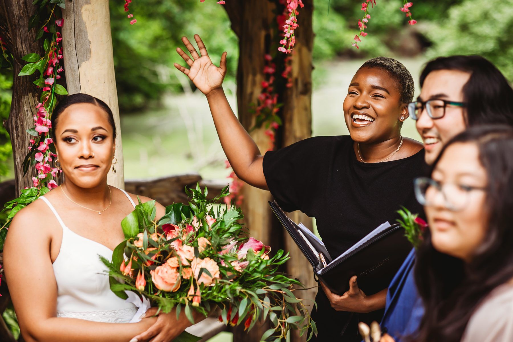 Robyn laughing with a bride and her guests beneath a rustic flowered arbor in Central Park
