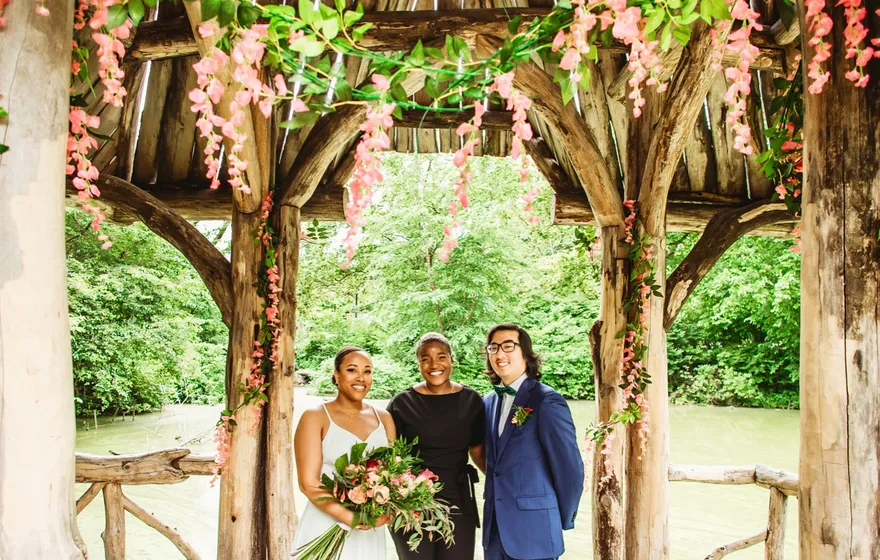 A bride with a bouquet, a groom in a blue suit and their officiant stand under the rustic timber shelter at Wagner Cove, flowers hung from the beams and the Lake behind them