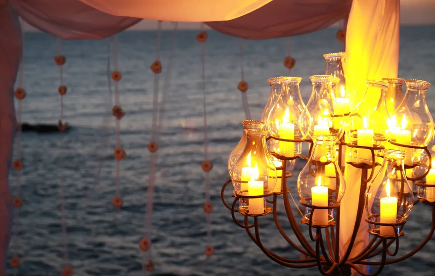 A tall candelabra of pillar candles, each sealed inside its own glass hurricane chimney, glowing gold beside a draped fabric arch at a seaside wedding at dusk