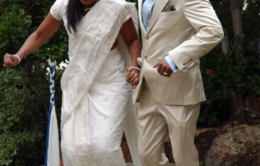 A bride in a white sari and a groom in a cream suit holding hands and jumping together over a broom resting on a small stand in a garden, both mid-air with their feet off the ground