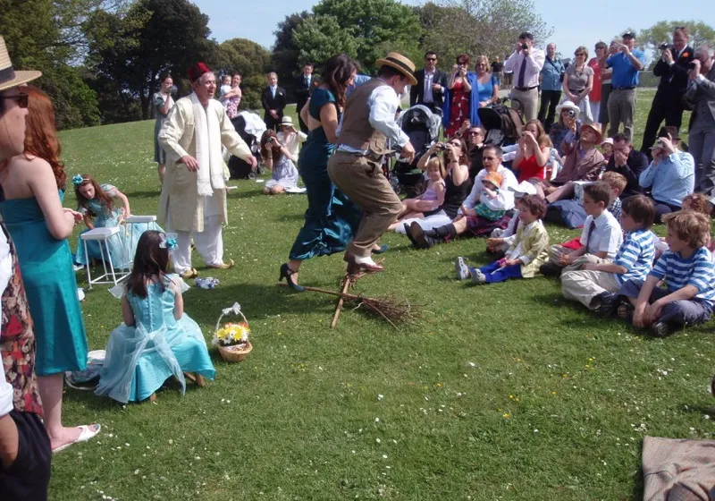 A couple in vintage dress jumping together over a broom laid on green grass at an outdoor wedding, friends and children seated in a circle watching and clapping in the sun