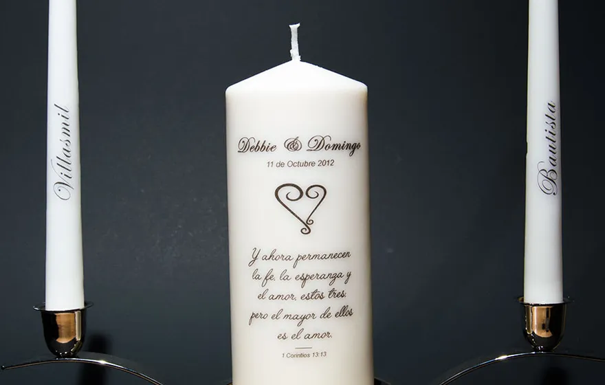 A studio shot of a unity candle set: an ivory pillar printed with the couple's names, date, and a verse, flanked by two named tapers standing in silver holders against a dark background
