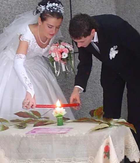 A bride in a lace gown and a groom in a dark suit lean in together to light a red pillar candle standing on a lace-covered table at their wedding