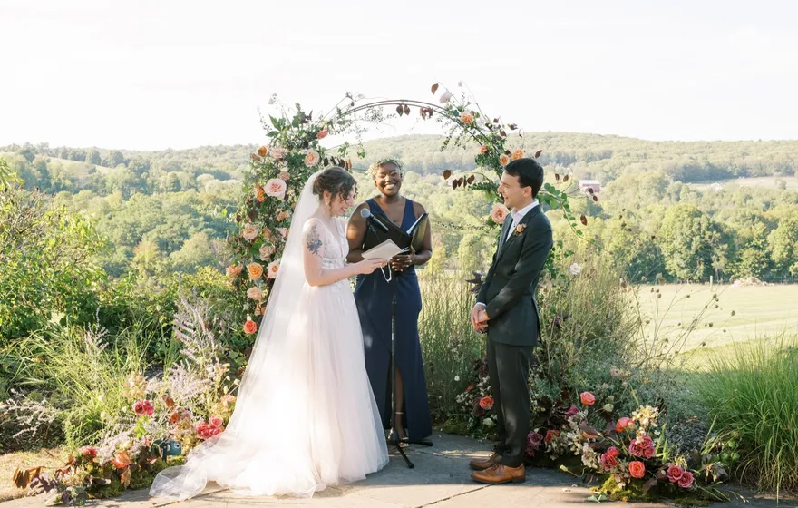 An officiant reads from a page at a flower-covered arch on a hilltop while the bride and groom face each other, green hills behind them