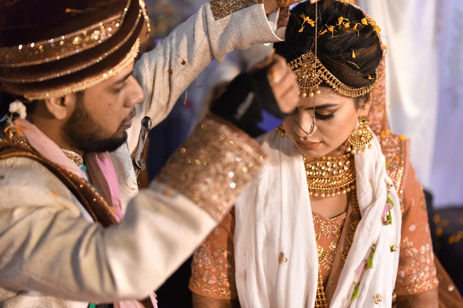 A groom in a sherwani and turban reaches over the bride during a Hindu wedding ritual, the bride in gold bridal jewelry and an embroidered lehenga