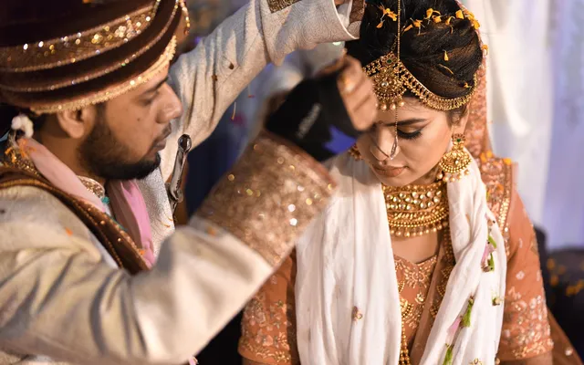A groom in a sherwani and turban reaches over the bride during a Hindu wedding ritual, the bride in gold bridal jewelry and an embroidered lehenga