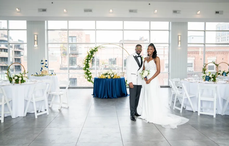 A Black bride and groom standing together in a bright loft reception space with floor-to-ceiling windows, white round tables and a navy sweetheart table with a floral arch behind them