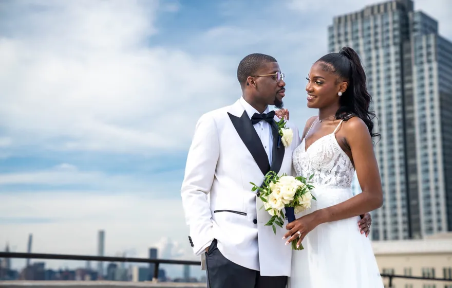 A Black couple on a sunlit rooftop, the groom in a white tuxedo jacket and the bride in a lace gown holding a white bouquet, gazing at each other with the city skyline behind them