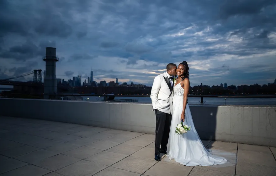 A Black bride in a white gown holding a bouquet and a groom in a white tuxedo jacket sharing a kiss on a Brooklyn rooftop at dusk, the Manhattan skyline and Williamsburg Bridge glowing behind them
