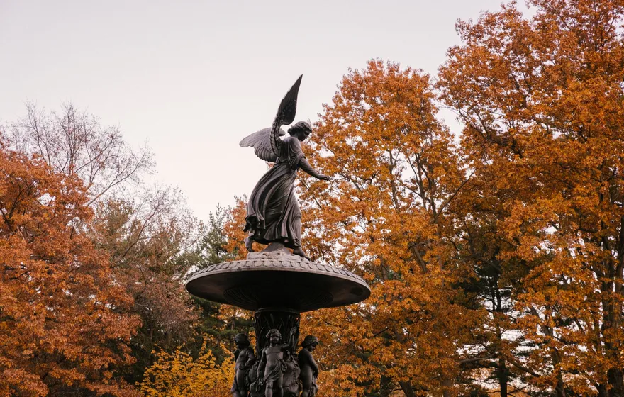 The bronze Angel of the Waters statue atop Bethesda Fountain, wings raised, against orange and red autumn trees